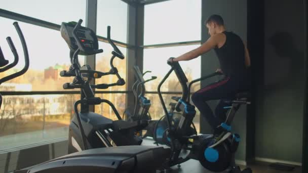 Jovem homem ativo girando uma bicicleta de ar no ginásio com treinadores. treinamento masculino em bicicleta de ar — Vídeo de Stock