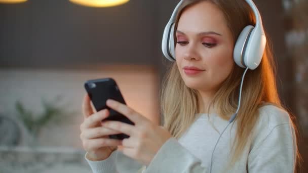 Calma mujer joven feliz en los auriculares escalofriante sentado en el sofá con los ojos cerrados escuchando música favorita sosteniendo el teléfono con aplicación de reproductor en línea móvil disfrutar de un estado de ánimo pacífico con auriculares — Vídeos de Stock