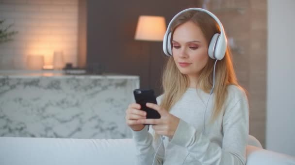Menina ouvindo música em fones de ouvido olhando para a tela do smartphone. Relaxe em casa — Vídeo de Stock