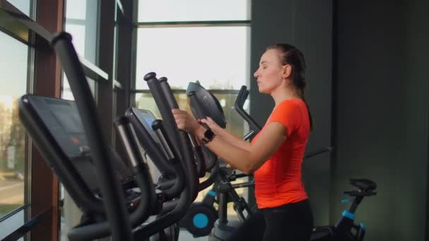 Belle forme sportive positive jeune femme dans le gymnase faire des exercices sur l'entraîneur elliptique de travail . — Video