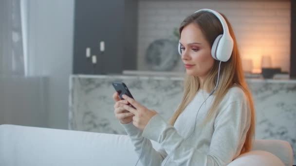 Chica joven escuchando música en auriculares mirando la pantalla del teléfono inteligente. Relájate en casa. — Vídeos de Stock