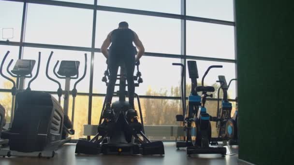 Vacker passform sportig positiv ung man i gym gör övningar på elliptisk tränare träna. Hårt — Stockvideo