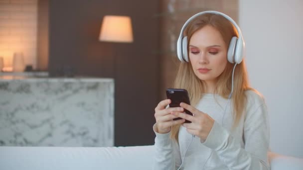Calm happy young woman in headphones chilling sitting on sofa with eyes closed listening to favorite music holding phone using mobile online player app enjoy peaceful mood wearing earphones — ストック動画