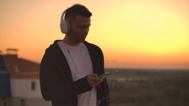 Büyük beyaz kulaklıklı bir adam gün batımında çatıda duran müzik dinliyor. Rahatlama