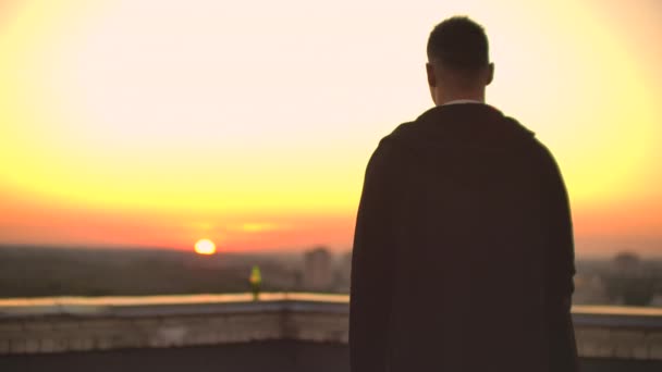 Tillbaka visa en manlig frilansprogrammerare i en hoodie promenader på taket vid solnedgången och dricker öl tittar på den vackra utsikten över staden — Stockvideo