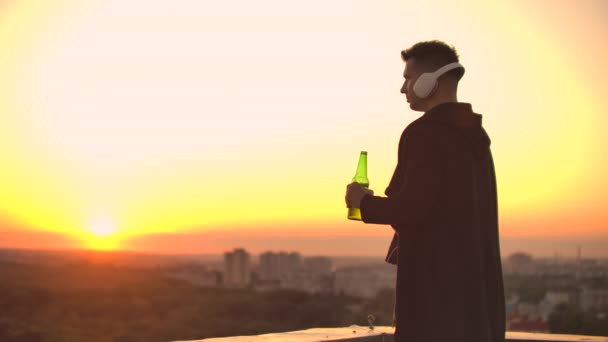 Чоловік п'є пиво, що стоїть на даху і думає про світ. Подивіться на вигляд міста на заході сонця з висоти. Дах хмарочоса — стокове відео