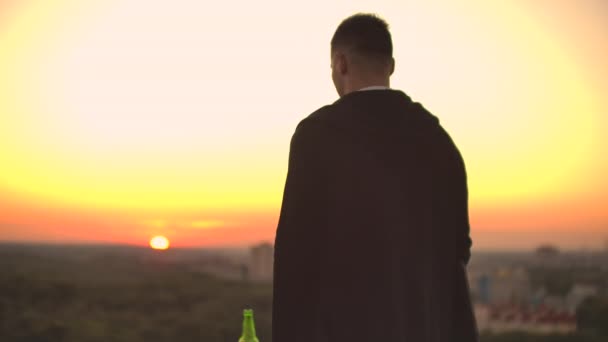 Vista desde atrás un hombre camina en el techo al atardecer y bebe cerveza mirando la hermosa vista de la ciudad — Vídeos de Stock