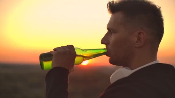 Close up de um homem bebendo cerveja ao pôr do sol em pé no telhado de um edifício contra o fundo de uma bela cidade à noite — Vídeo de Stock