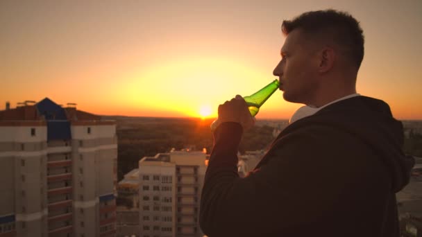 Ένας άντρας πίνει μπύρα στην ταράτσα και σκέφτεται τον κόσμο. Κοιτάξτε τη θέα της πόλης στο ηλιοβασίλεμα από ύψος. Η οροφή ενός ουρανοξύστη — Αρχείο Βίντεο