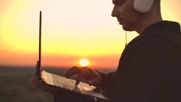 A man in large white headphones types with his fingers on the keyboard of a laptop standing on the roof of a building at sunset against the background of the city — Stock Video