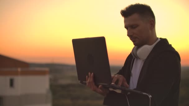 De pie al atardecer en el techo con un ordenador portátil y una cerveza. Un hombre en una sudadera con capucha trabaja divirtiéndose y contemplando la belleza de la vista de la ciudad desde una altura — Vídeo de stock