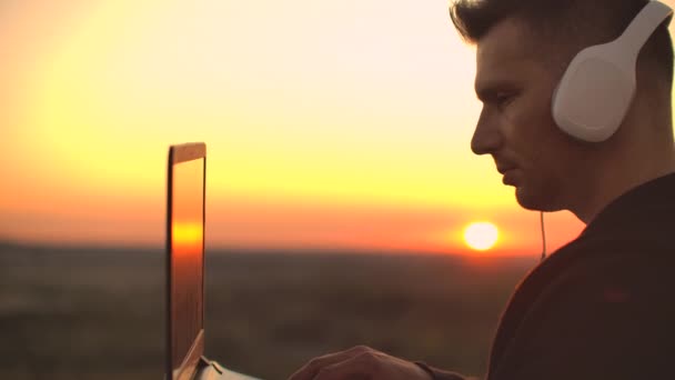 Debout au coucher du soleil sur le toit avec un ordinateur portable et de la bière dans les écouteurs fonctionnent à distance et contempler la beauté. Être en harmonie fonctionne à distance avec votre ordinateur portable — Video