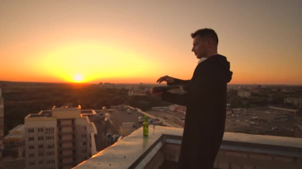 Schließen Sie den Deckel des Laptops und trinken Sie Bier stehend auf dem Dach. ein Mann im Kapuzenpullover mit einem Laptop in der Hand bei Sonnenuntergang — Stockvideo