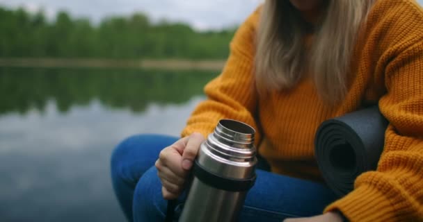 Una viaggiatrice seduta sul molo di legno del lago in un maglione giallo con uno zaino che beve tè caldo o caffè da un thermos guardando la bellezza della natura. Godersi il momento — Video Stock