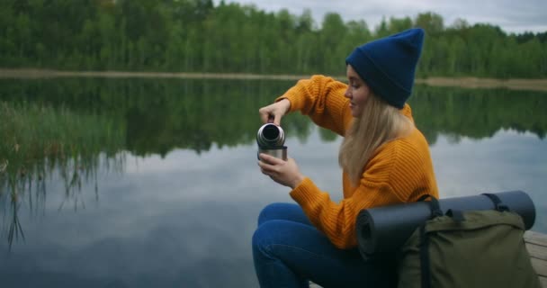 Eine Reisende sitzt in einem gelben Pullover und einem Rucksack auf dem Holzsteg des Sees und trinkt heißen Tee oder Kaffee aus einer Thermoskanne und betrachtet die Schönheit der Natur. Den Moment genießen — Stockvideo