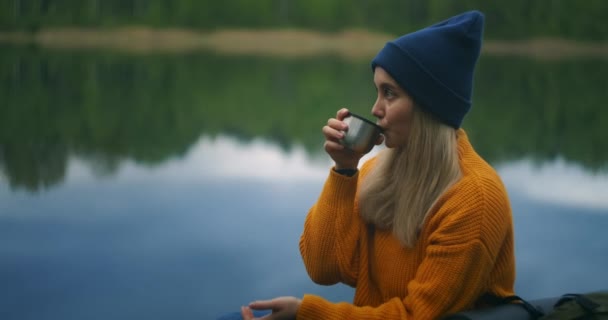 女游客穿着黄色毛衣坐在湖边的木制码头上，背着背包，喝着热茶或热水瓶中的咖啡，凝视着大自然的美丽。享受这一刻 — 图库视频影像