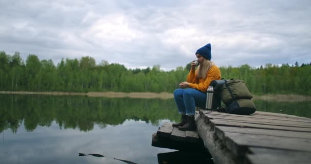 Uma viajante feminina sentada no cais de madeira do lago em uma camisola amarela com uma mochila bebendo chá quente ou café de uma garrafa térmica olhando para a beleza da natureza. Aproveitando o momento — Vídeo de Stock
