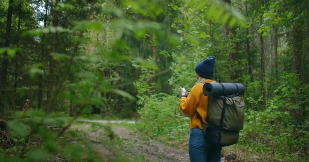 Cámara lenta: Joven excursionista usando su teléfono inteligente mientras disfruta de su fin de semana en el bosque. Mujer viajera con mochila caminando por el camino del bosque mirando el teléfono y el mapa en madera verde — Vídeo de stock