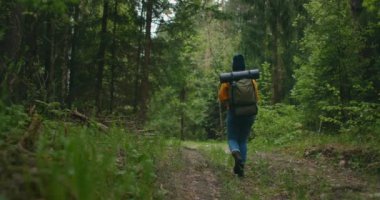 Genç bir kadın sırtında sırt çantasıyla ormanda bir yol boyunca ağır çekimde yürüyor. Sarı kazaklı ve şapkalı bir kadın gezgin sonbaharda ya da baharda orman yolunda yürüyor..
