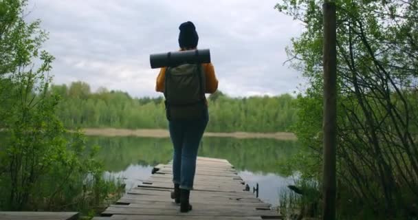 Blick von hinten: Eine Frau mit Rucksack geht über eine Holzbrücke durch den Wald zum See — Stockvideo