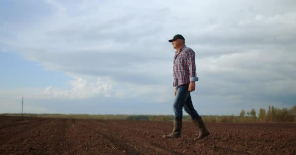 Movimento lento: macho sênior um motorista de trator caminha através de um campo arado ao pôr do sol após o trabalho — Vídeo de Stock