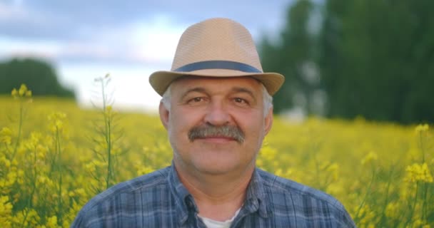 Κοντινό πλάνο του πρεσβύτερου Καυκάσου Κοιτάζοντας την κάμερα ένας όμορφος σοφός αγρότης με καπέλο κοιτάζει στο πλάι, γυρνώντας το πρόσωπο στην κάμερα και χαμογελώντας στο χωράφι με τους σπόρους. Πορτρέτο — Αρχείο Βίντεο