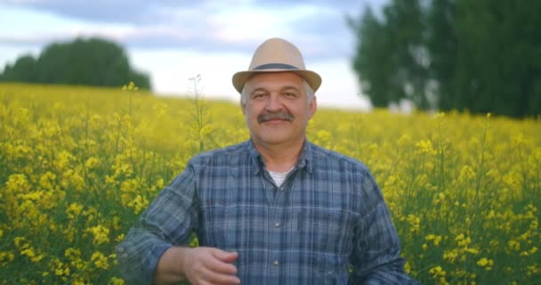 Primer plano de la persona mayor del Cáucaso Mirando a la cámara de hombre sabio bien parecido granjero en un sombrero mirando a un lado, girando la cara a la cámara y sonriendo en el campo de colza de trigo. Retrato — Vídeo de stock