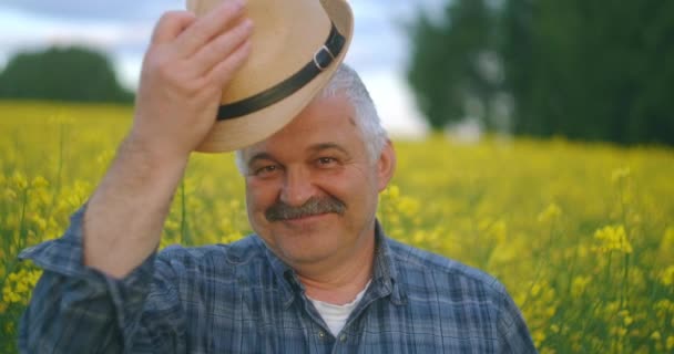 Primer plano de la persona mayor del Cáucaso Mirando a la cámara de hombre sabio bien parecido granjero en un sombrero mirando a un lado, girando la cara a la cámara y sonriendo en el campo de colza de trigo. Retrato — Vídeo de stock