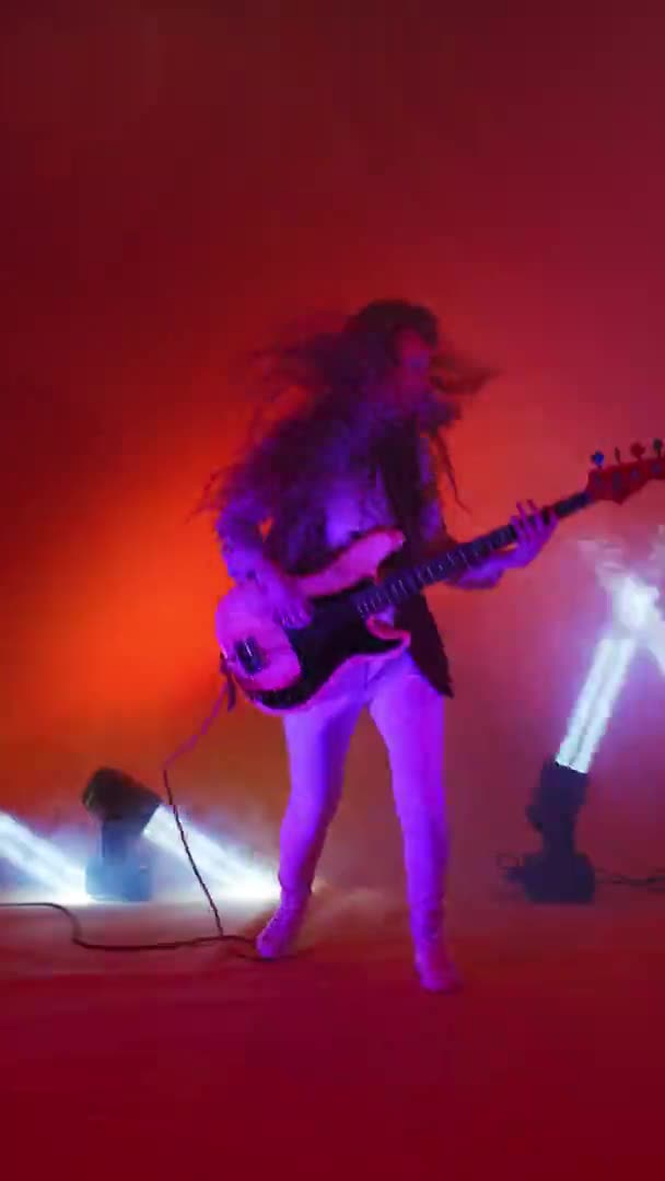 Verticale video van een vrouwelijke bassist in een jasje die op één been danst en haar hoofd en haar schudt. Gek gitarist in de studio neon lichten en spots — Stockvideo