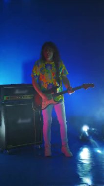 Renkli ışıklı dumanlarda zıplayan ve gitar çalan dikey video, neşeli adam. Stüdyoda değişen bir arka planda sahne ışıkları.