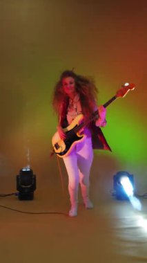 Ceketli, tek bacaklı, dans eden, başını ve saçlarını sallayan kadın basçının dikey videosu. Stüdyo neon ışıkları ve spot ışıkları altında çılgın bir gitarist.
