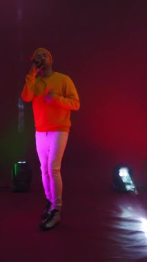 Dikey video. Komik erkek rap sanatçısı bir mikrofona şarkı söylüyor ve dans ediyor lambaların neon ışığında ve stüdyoda renkli parlak bir arka planda.