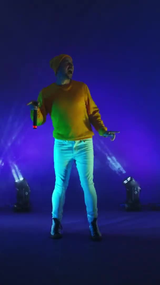 Vertical video. Incendiary funny Singer in yellow clothes dancing and singing with a microphone in neon color. Jump and move vigorously. Hip-hop and pop artist — Stock Video