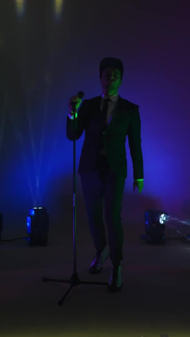 Dikey video. Ceketli şık bir adam mikrofon standında dans ediyor ve stüdyoda neon ışıkta şarkı söylüyor. Enerjik ve etkileyici bir şarkıcı. — Stok video