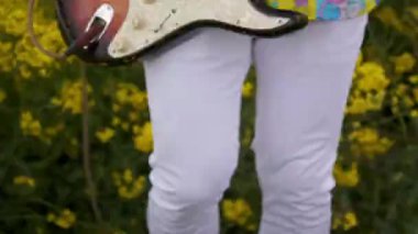 Kameranın manuel hareketi dans eden gitaristi takip ediyor. Tişörtlü bir adam, kolza tarlasında gitar çalıyor..