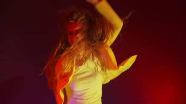 En rolig kvinna i gul jacka hoppar och dansar energiskt i neonljuset och röker — Stockvideo