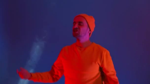 Muž vesele tančí a dívá se do kamery ve světle zářivých světel a barevných reflektorů. Neonové barvy a bláznivý muž tančí na hudbu a zpívá ve žluté bundě a klobouku — Stock video