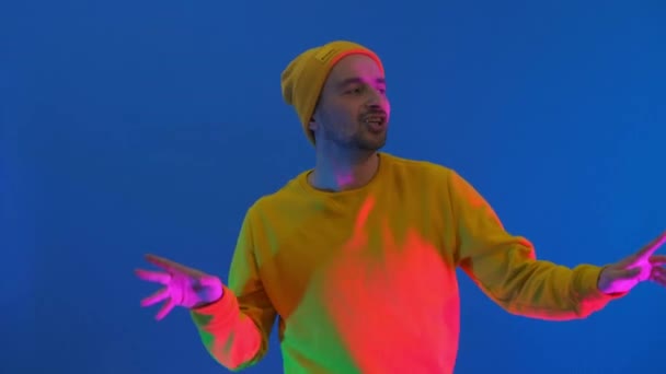 Un homme avec un sweat à capuche jaune et un chapeau danse sur fond bleu. Danse drôle en regardant la caméra