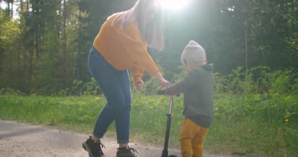 Una joven madre le enseña a su hijo a montar en un scooter apoyándolo y animándolo. Madres amor y cuidado . — Vídeos de Stock