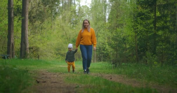 Egy fiatal anya együtt sétál a parkban egy gyerekkel, aki fogja a kezét. Együtt sétáltunk az erdőben. Anyaság és gyermekkor. Anya elkíséri a gyermeket, amikor az erdőben sétál. — Stock videók