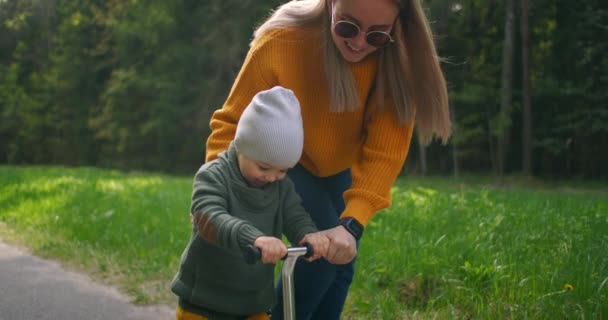 Retrato de un niño feliz montando una moto con su madre en el parque. un niño aprende a montar un scooter — Vídeos de Stock