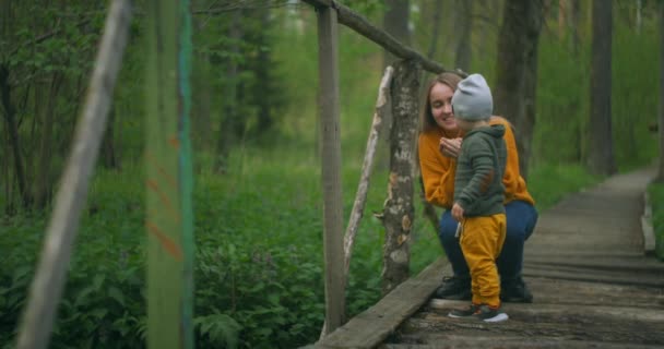 Na moście leśnym matka i syn wrzucają kamienie do strumienia. Matka i chłopiec spędzają razem czas na spacerach po parku — Wideo stockowe