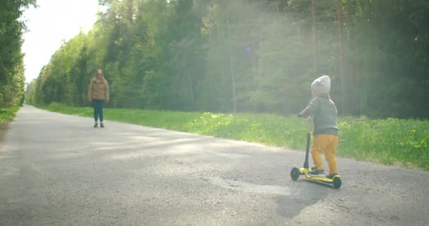 公園で彼のお母さんと一緒に男の子はスクーターに乗ると男の子はスローモーションで彼のお母さんに会うために乗る — ストック動画