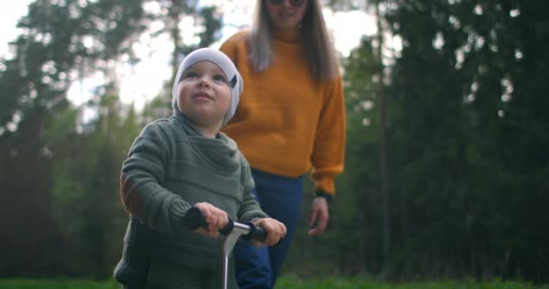 Ralenti un garçon souriant heureux chevauchant un scooter dans le parc avec sa mère. — Video
