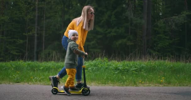 Vista lateral, a mãe ajuda o menino com o desenvolvimento de uma scooter. Ensinar o meu filho a andar de scooter. Maternidade e infância — Vídeo de Stock