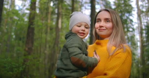 Μια νεαρή μητέρα περπατά μαζί στο πάρκο με ένα παιδί να το κρατάει στην αγκαλιά της. Αγκαλιάστε το γιο σας και περπατήστε μαζί στο δάσος. — Αρχείο Βίντεο