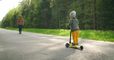 Çocuk scooter sürmeyi öğreniyor ve ağır çekimde annesiyle buluşmaya gidiyor yolda bir scooterla çam ağaçlarıyla dolu bir parkta.