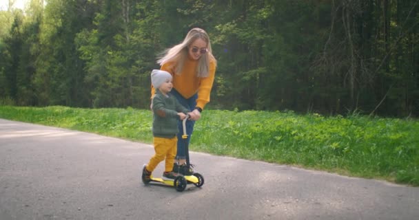 O menino aprende a andar de scooter e em câmera lenta. A mãe ajuda o rapaz no treino. O conceito de apoio e apoio às crianças . — Vídeo de Stock