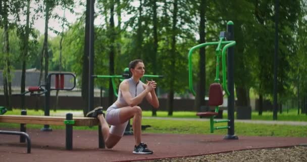 En ung vacker kvinna ensam utför övningar i parken på sommaren på en bänk. Hälsosam livsstil och idrottsträning i parken på sommaren. — Stockvideo