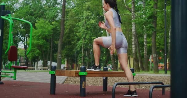Una joven en un parque realiza saltos a pie en un banco en ropa deportiva en el verano. Atletismo Mujer caucásica entrena en el Parque . — Vídeos de Stock